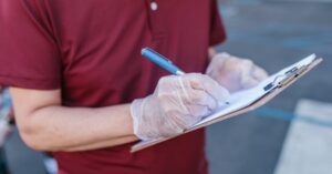 A person wearing gloves writes on a clipboard outdoors in casual attire.