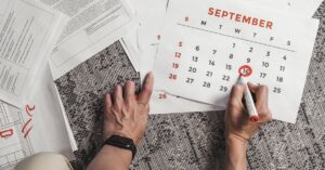 Hands holding a calendar with September dates encircled, surrounded by documents on a carpet.